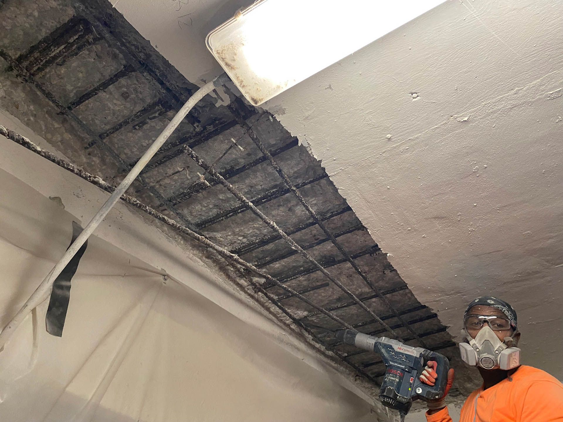 Man in respirator chipping away at concrete ceiling with jackhammer.