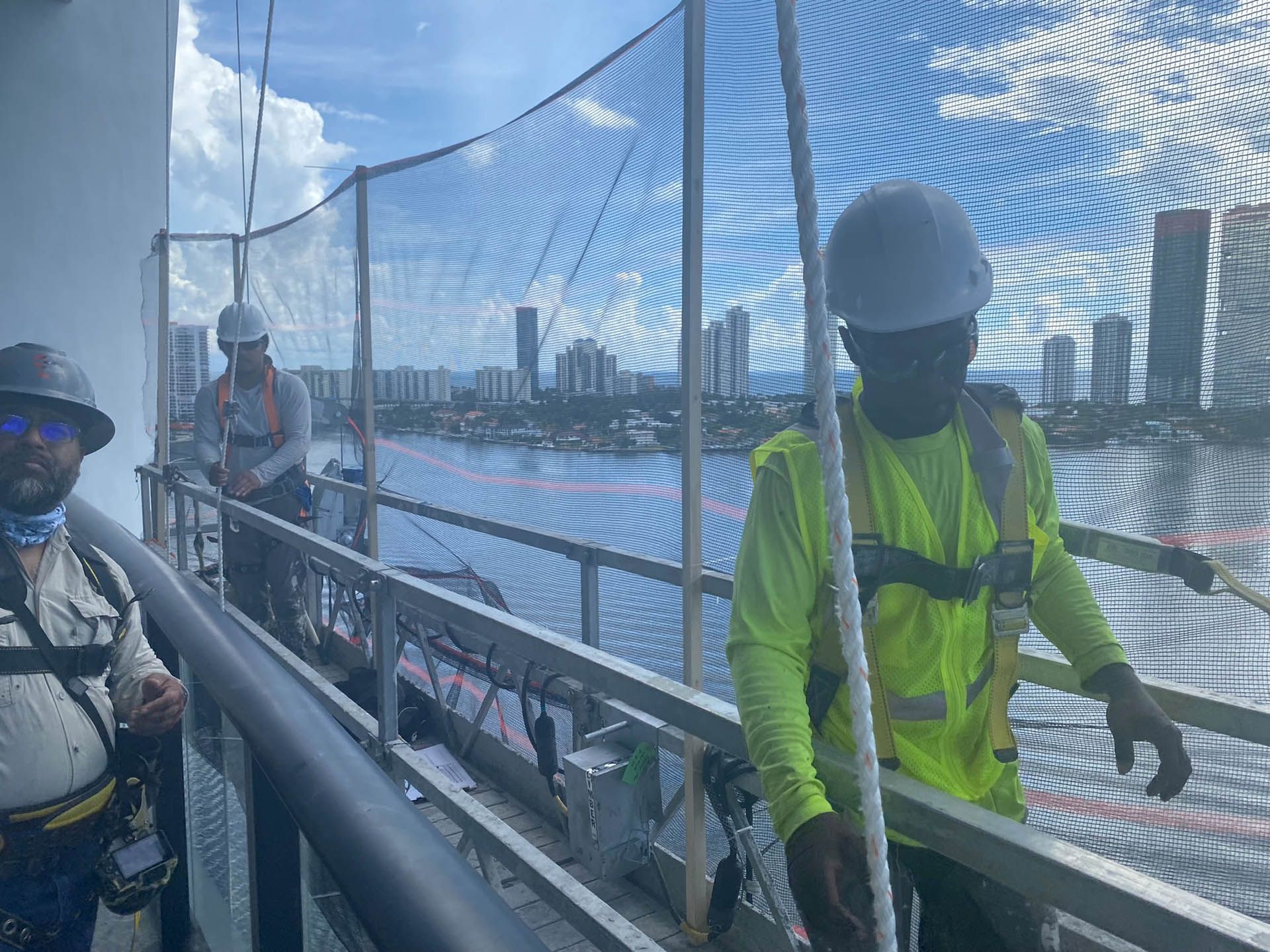 Workers on a high bridge, wearing safety gear and hard hats. Nets and city skyline in the background.