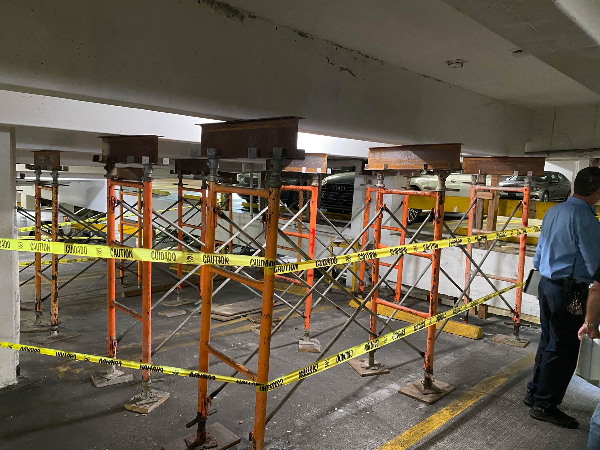 Orange scaffolding supports a parking garage ceiling; caution tape and a person are in view.