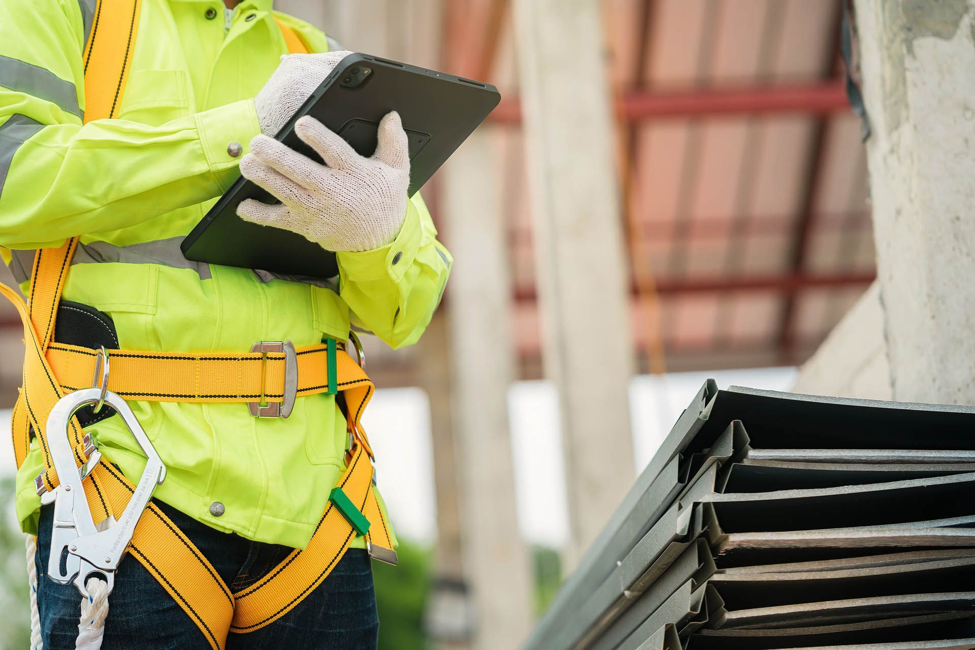 Worker in safety vest and harness holding a tablet at a construction site.