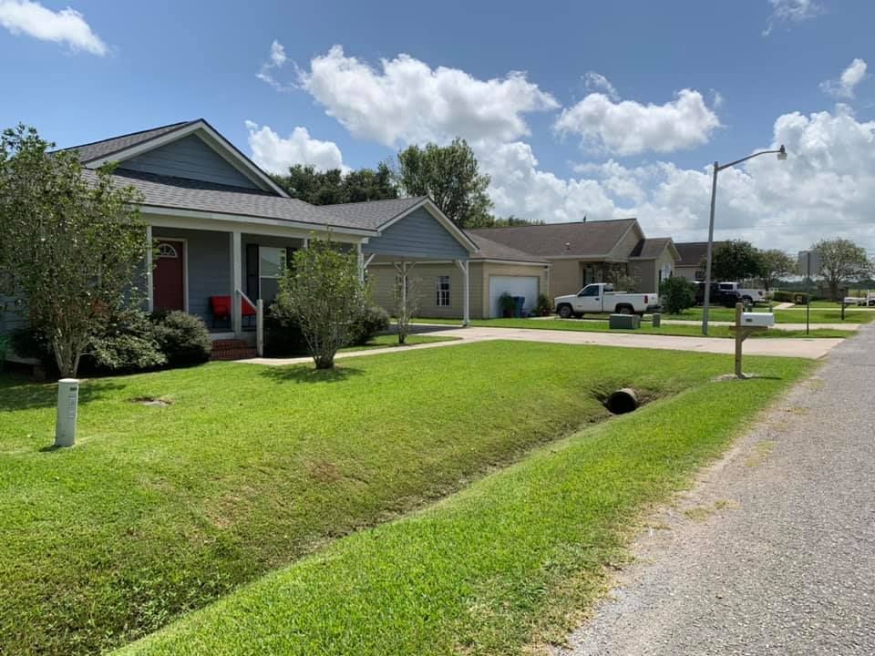 A suburban street with blue houses, mowed lawns, and a ditch in the foreground under a cloudy blue sky.