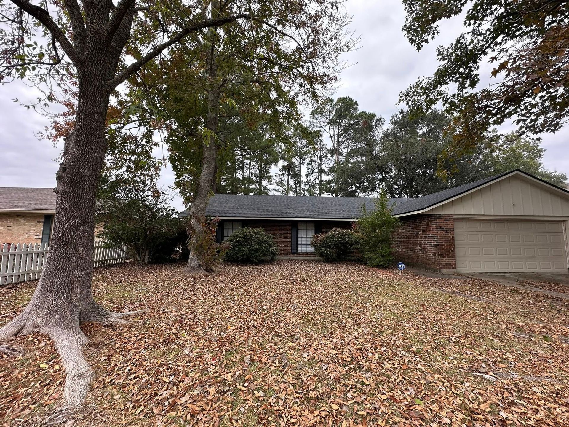 A single-story brick house with a two-car garage, surrounded by large trees and a yard covered in fallen autumn leaves.