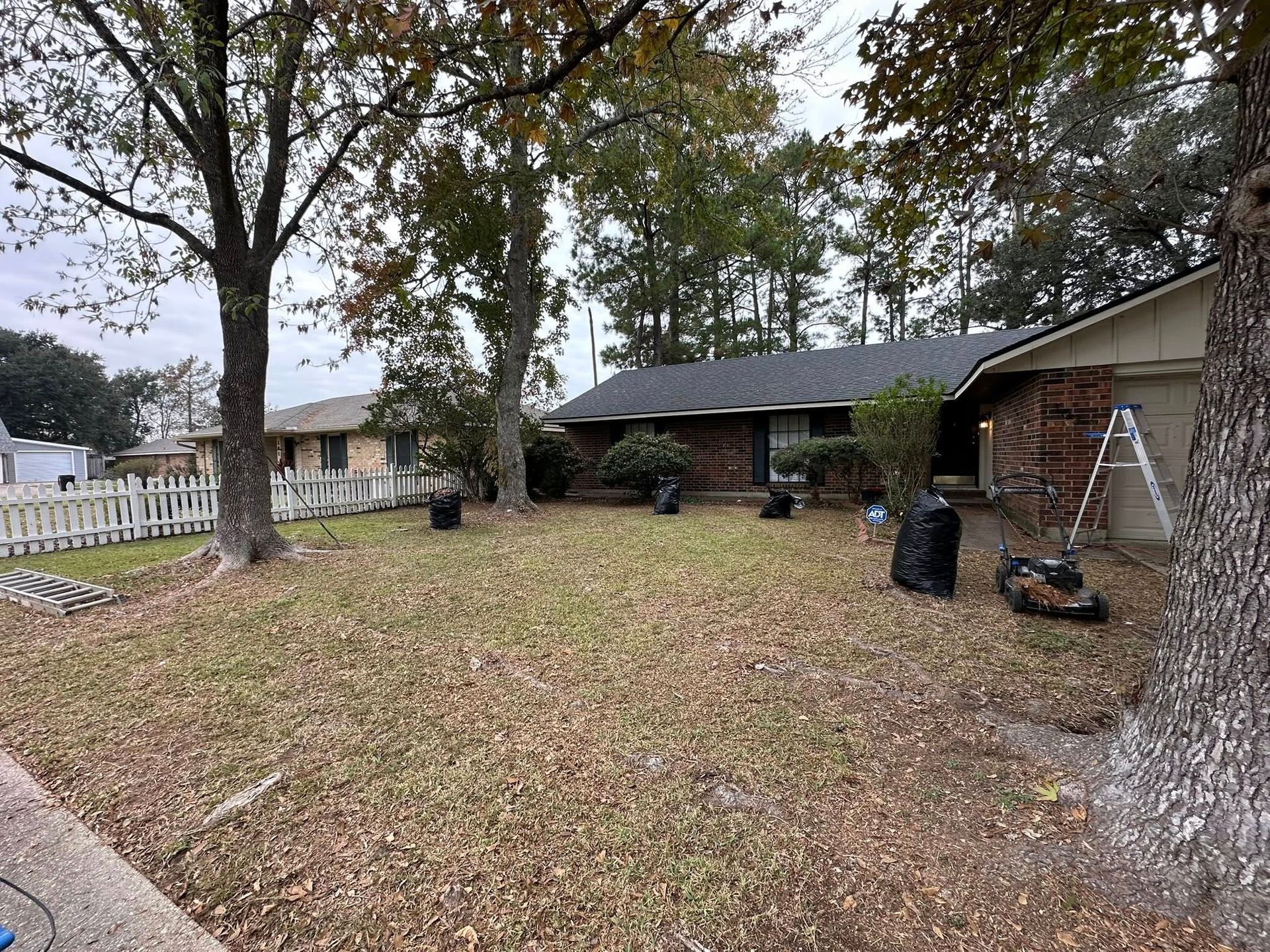 A house with a brick exterior, a grey roof, and a leaf-covered front lawn with several black yard waste bags.