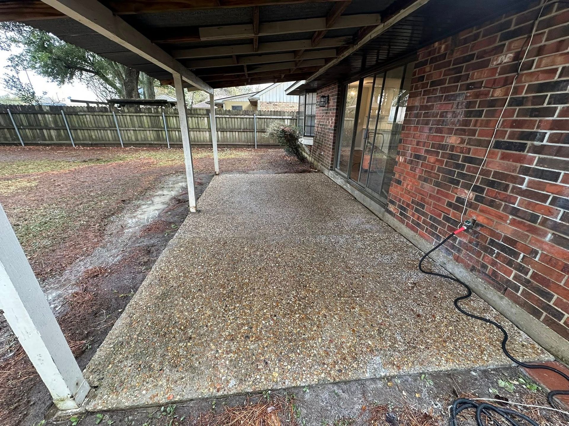A covered concrete patio with a pebbled texture adjacent to a red brick wall, overlooking a grassy yard with a fence.
