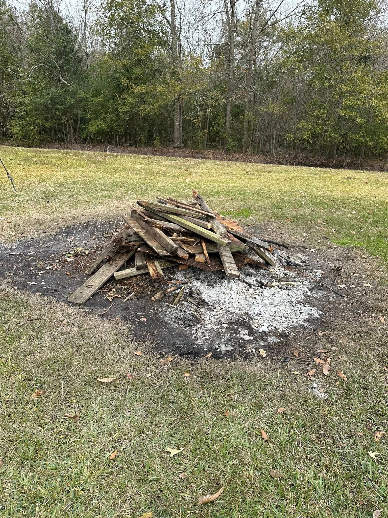 A pile of wood scraps and charred ash sits on a patch of dead grass in a wooded area.