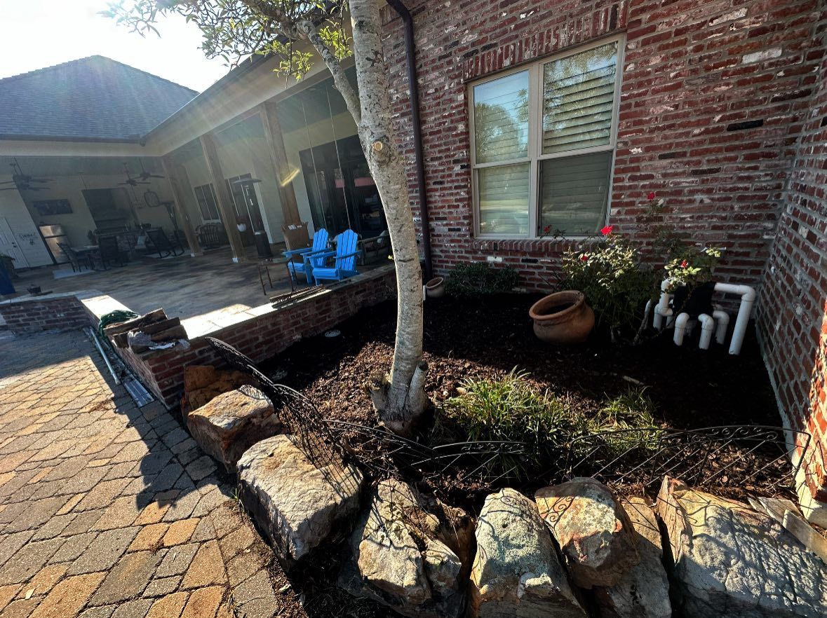 A brick home exterior with a garden bed, large landscape stones, a small tree, and blue chairs on a patio in the background.