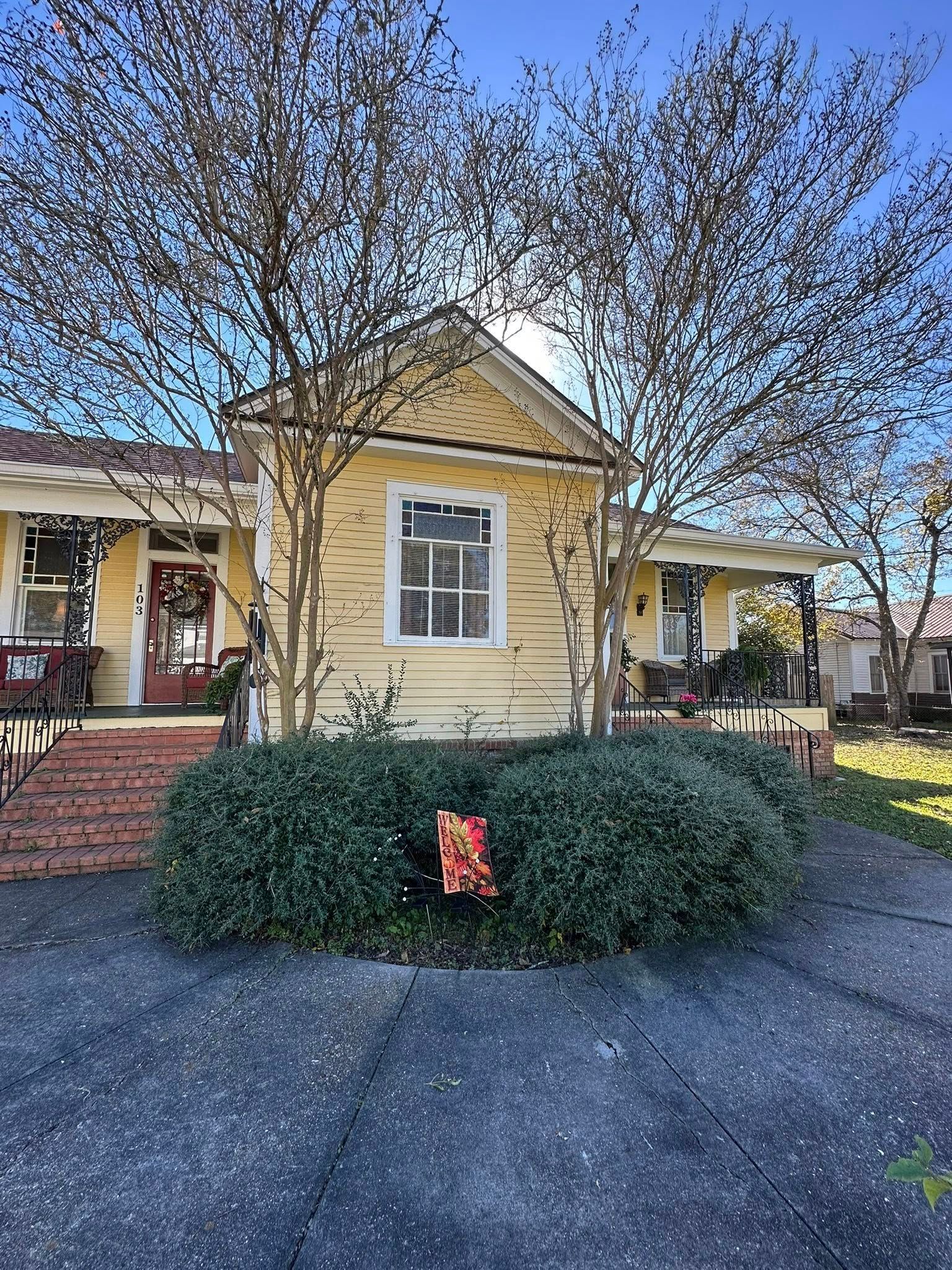 A yellow, one-story house with a white-trimmed window, a covered porch, and a large bush in the front yard.