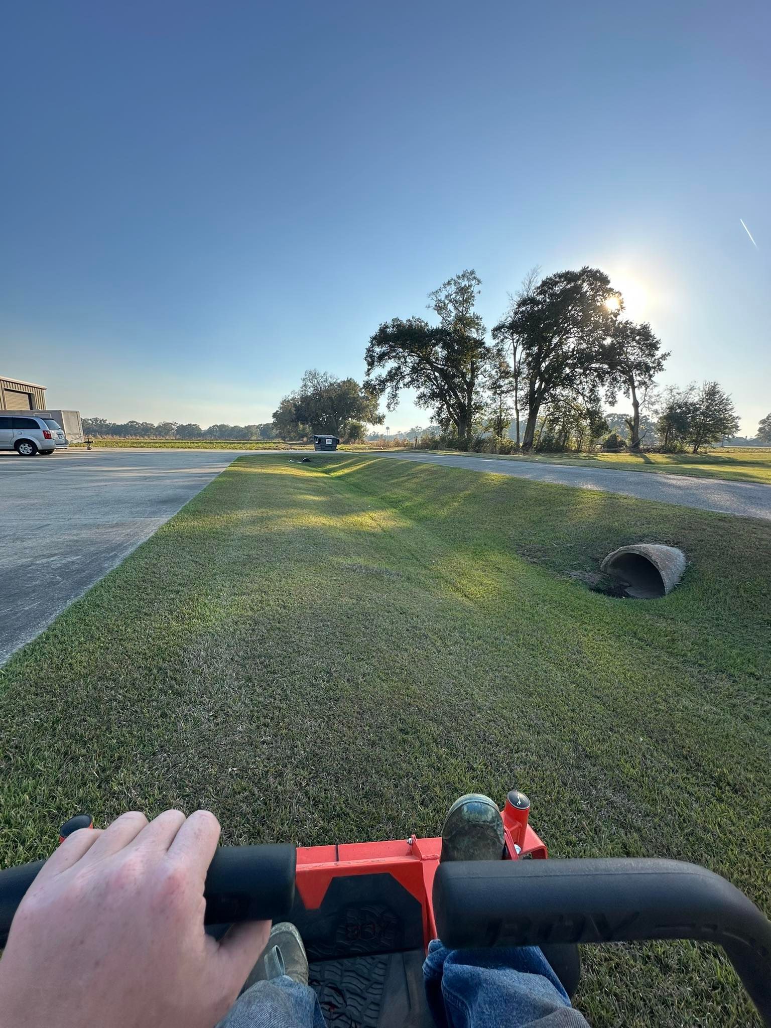 First-person view of someone operating a red lawnmower over grass, with a drainage pipe and trees in the background.