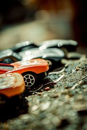 Toy cars lined up on gravel, including orange, silver, and black cars in shallow focus.