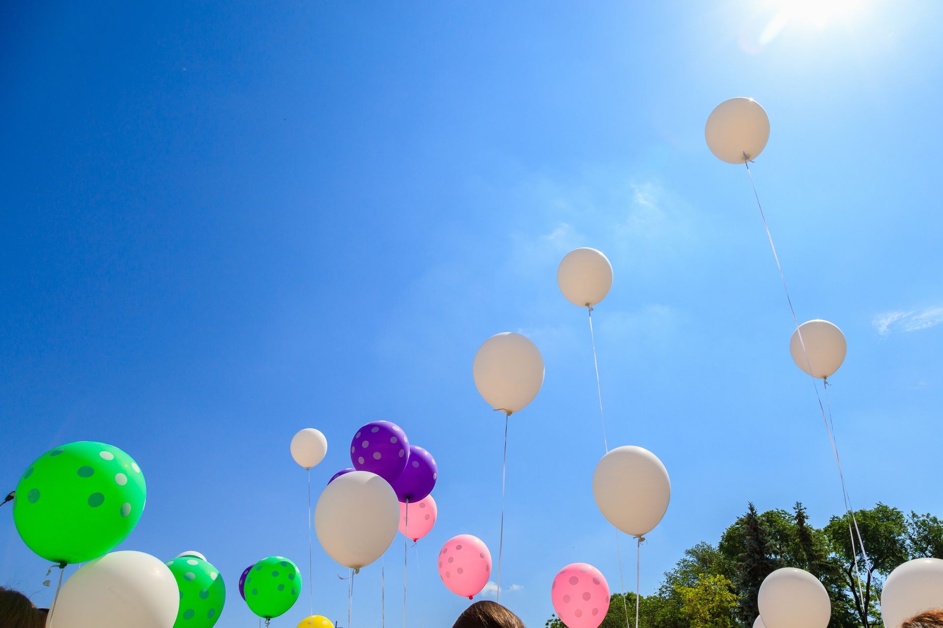 Balloons released into a bright blue sky. Mostly white balloons with pops of green, purple, and pink. Trees at bottom right.