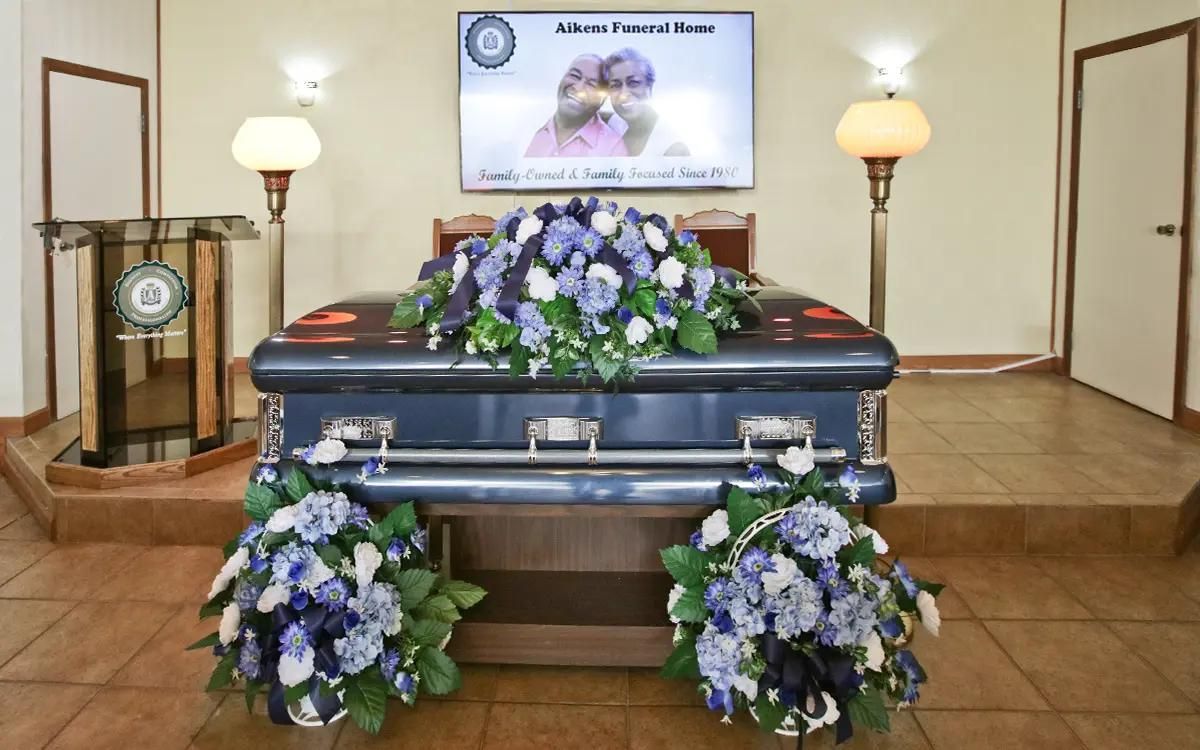 Blue casket with floral arrangements in a funeral home; screen displays a photo.
