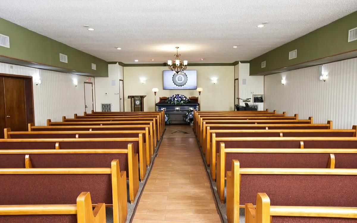 Interior of a funeral home chapel with rows of brown pews facing a flower-adorned casket.