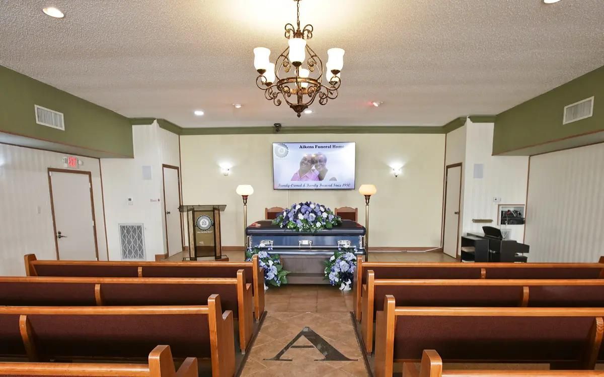 Interior of a funeral home chapel with rows of pews facing a casket draped in flowers, with a video screen above.