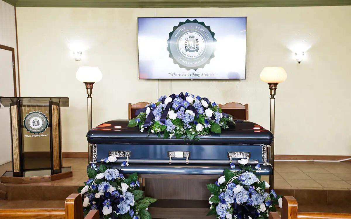 Blue casket adorned with blue and white flowers at a funeral, with a floral wreath on top and floral arrangements at the base.