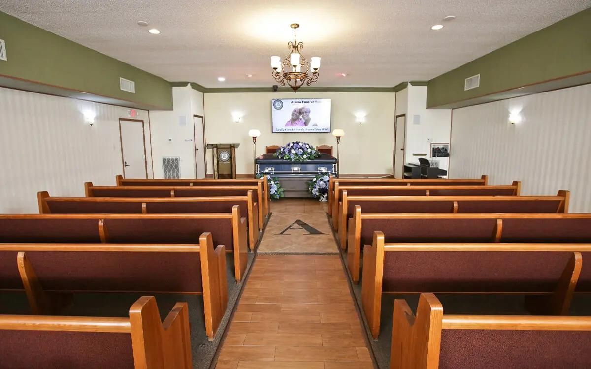 Funeral home chapel with rows of brown pews, a casket, and a television screen.