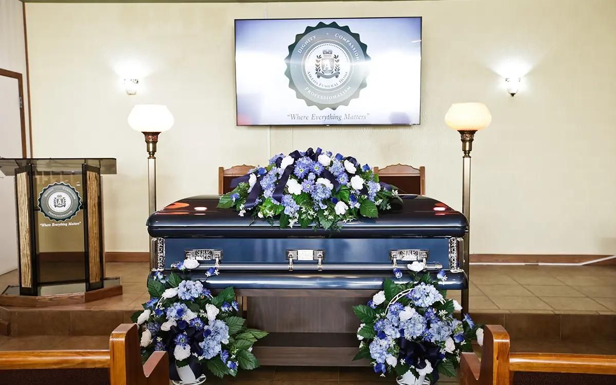 A dark blue casket with floral arrangements in a funeral home, television and lectern in the background.