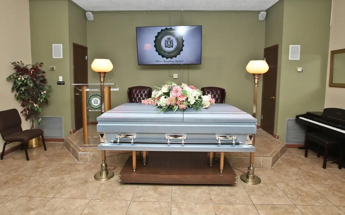 A casket adorned with flowers in a funeral home; two lamps and a piano are in view.