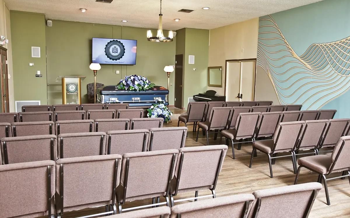 Funeral home chapel with rows of chairs facing a blue casket with floral arrangements. Green walls, a TV, and a decorative wall design.
