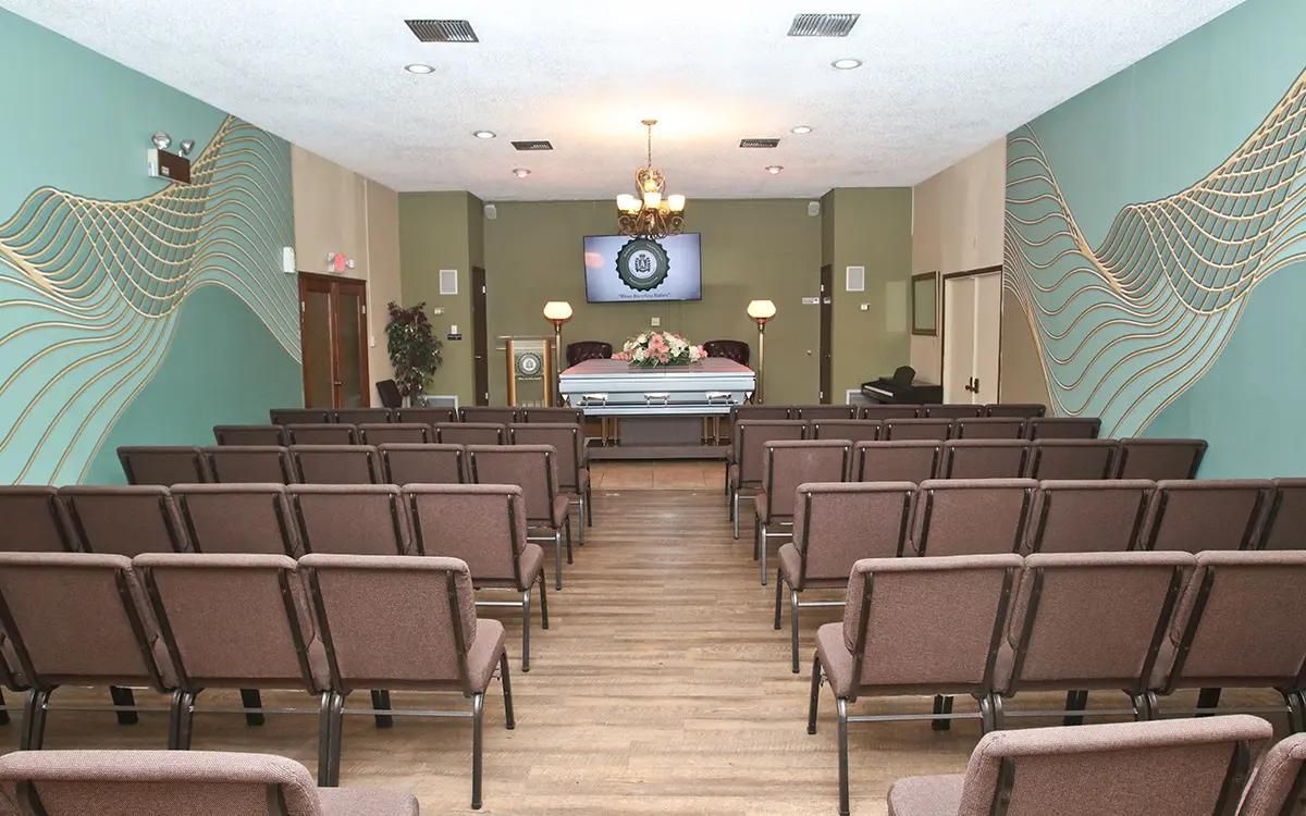 Interior of a funeral home chapel with rows of chairs facing a casket. Green and gold wall decor.
