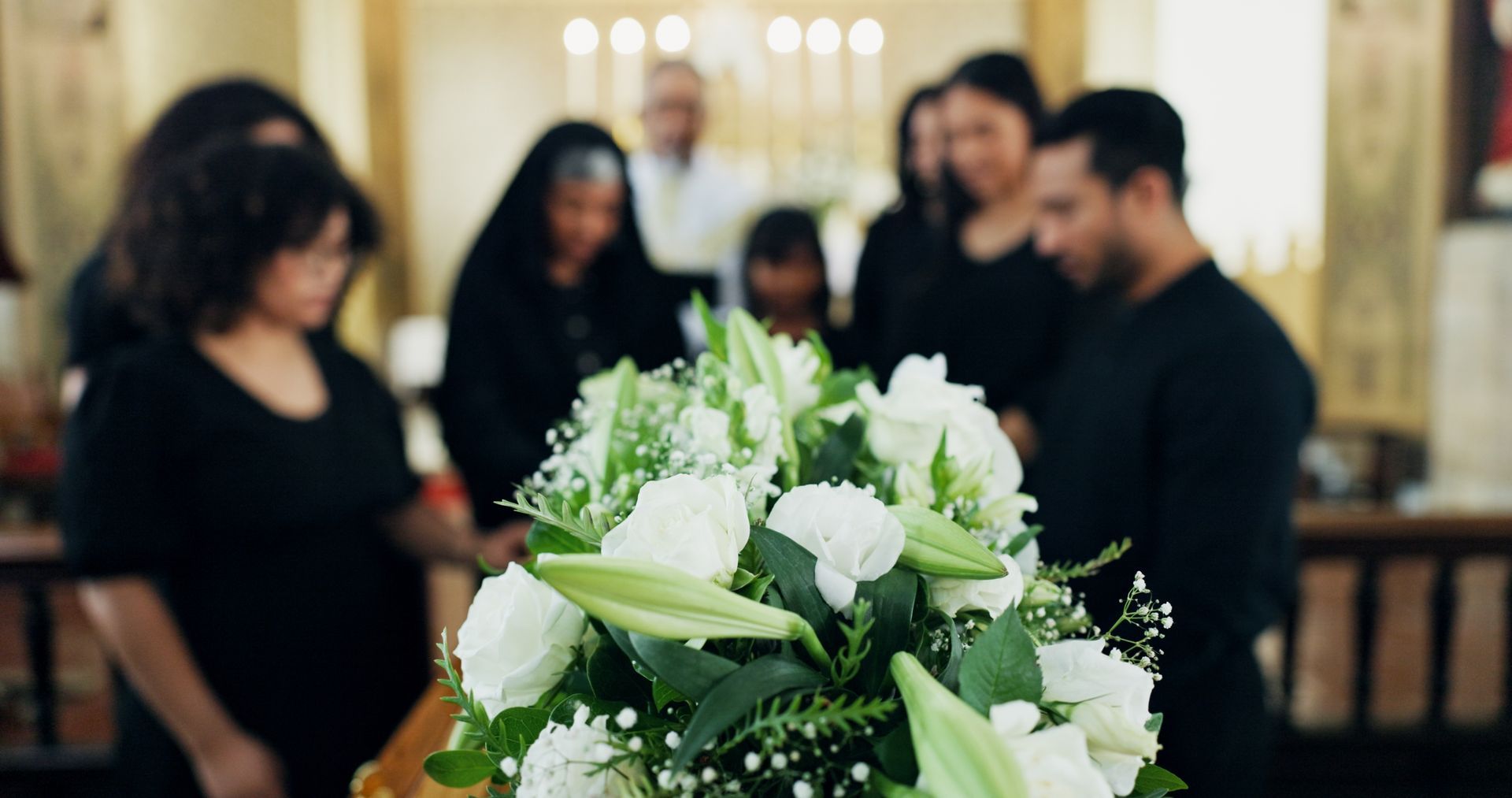 People at a funeral, gathered around a casket with white flower arrangement. They are somber, indoors.