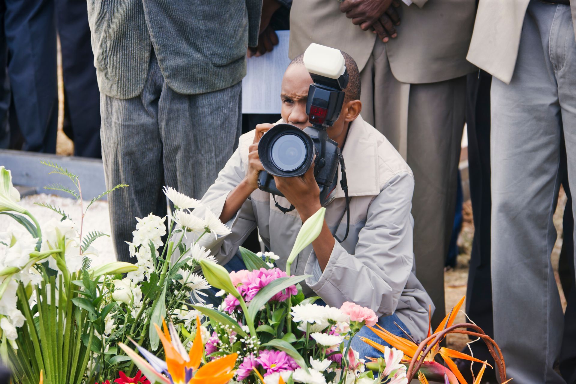 Photographer kneels near flowers, taking a photo with a camera. People in suits stand in the background.