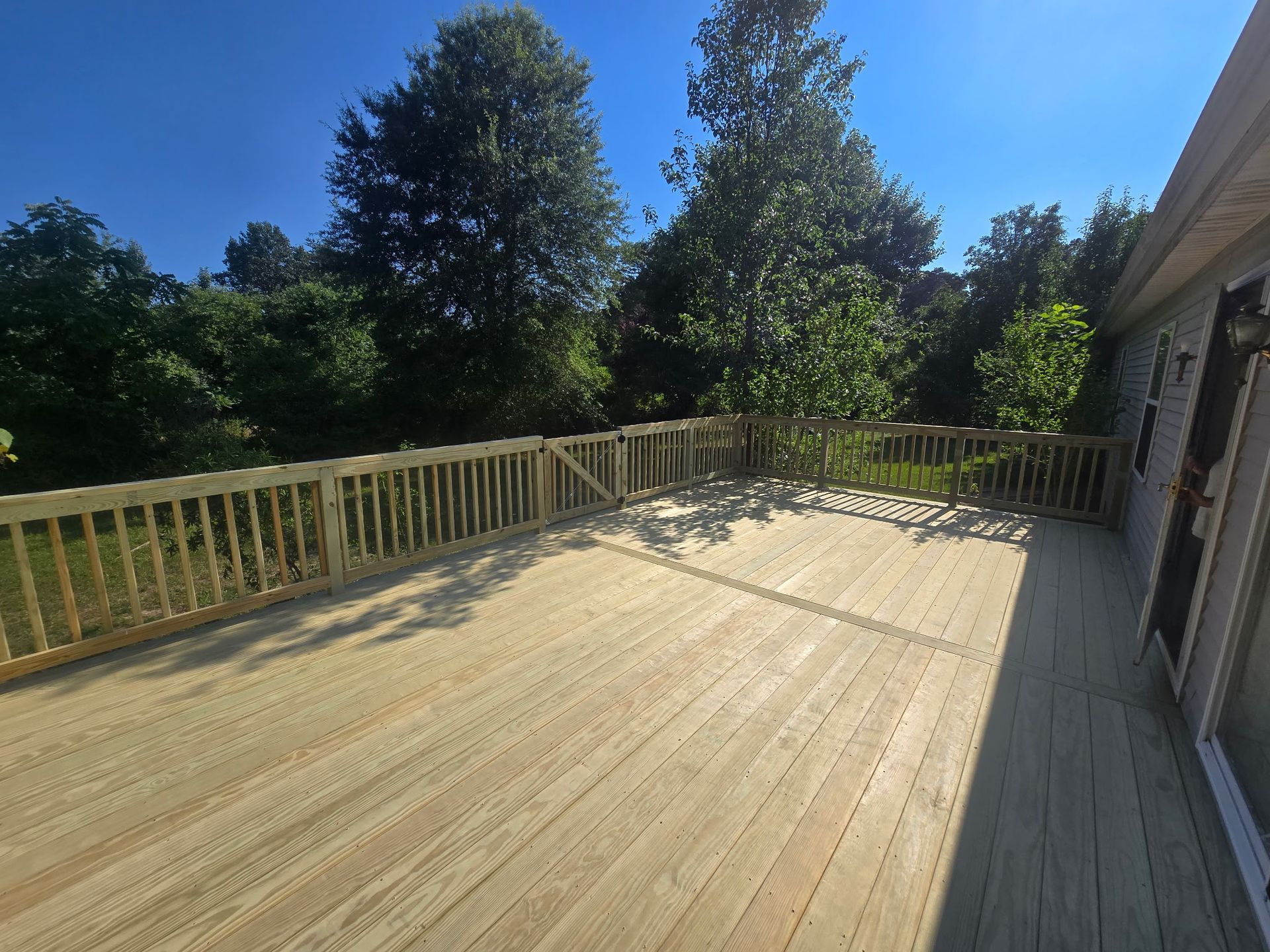 Sunny wooden deck with railing beside a house, overlooking trees and blue sky