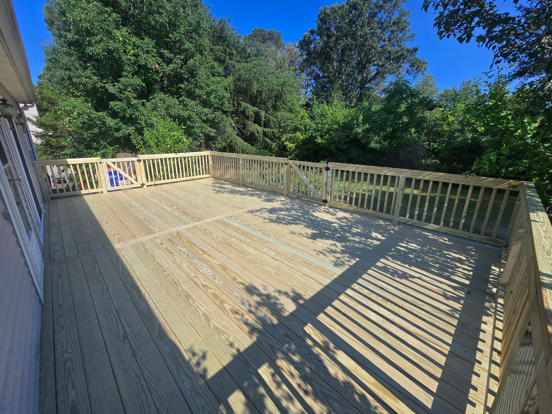 Wooden deck with railing in bright sunlight, surrounded by trees and blue sky