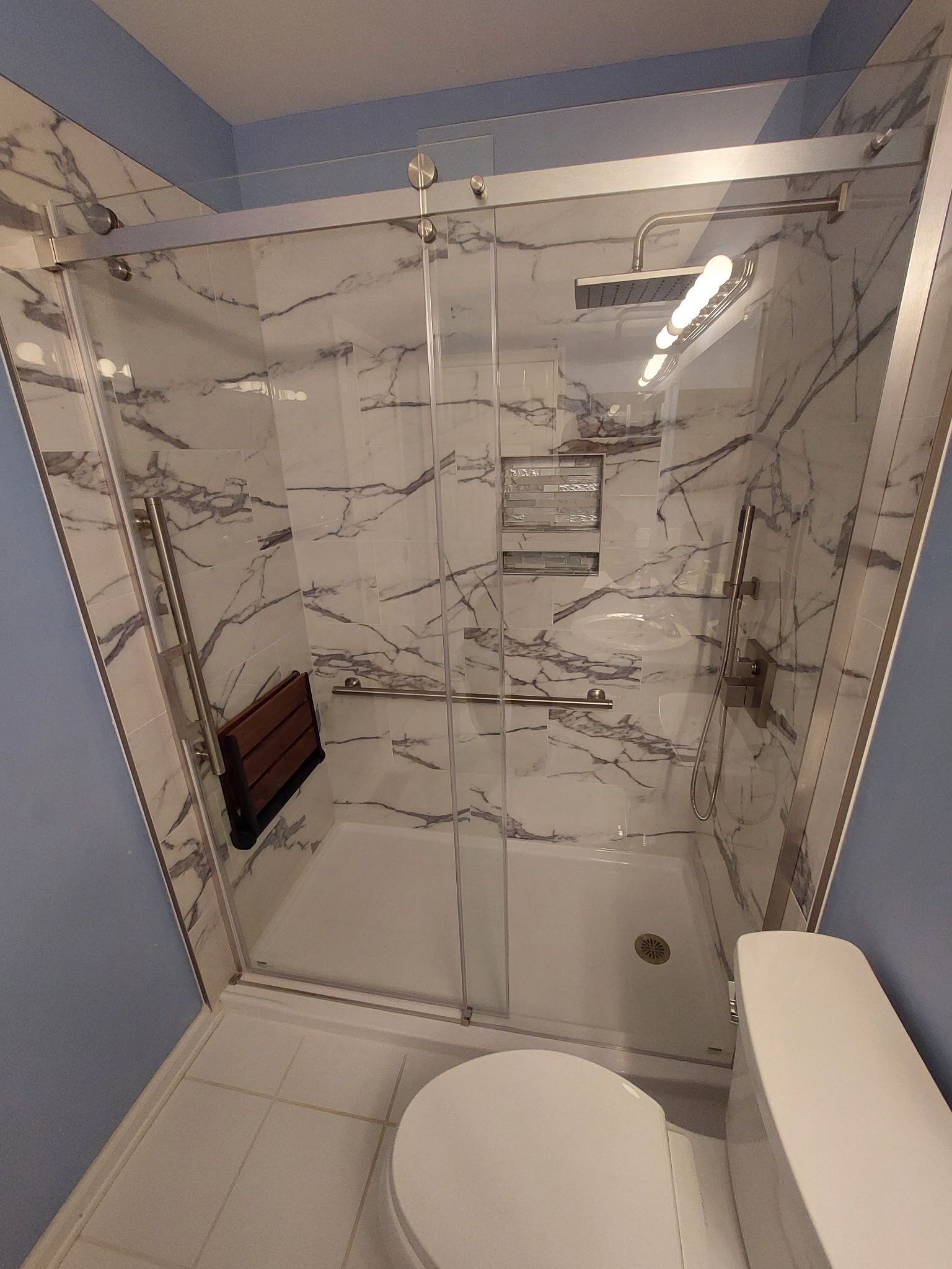 Modern marble-tiled shower with glass door, wall niche, wooden bench, and toilet in the foreground