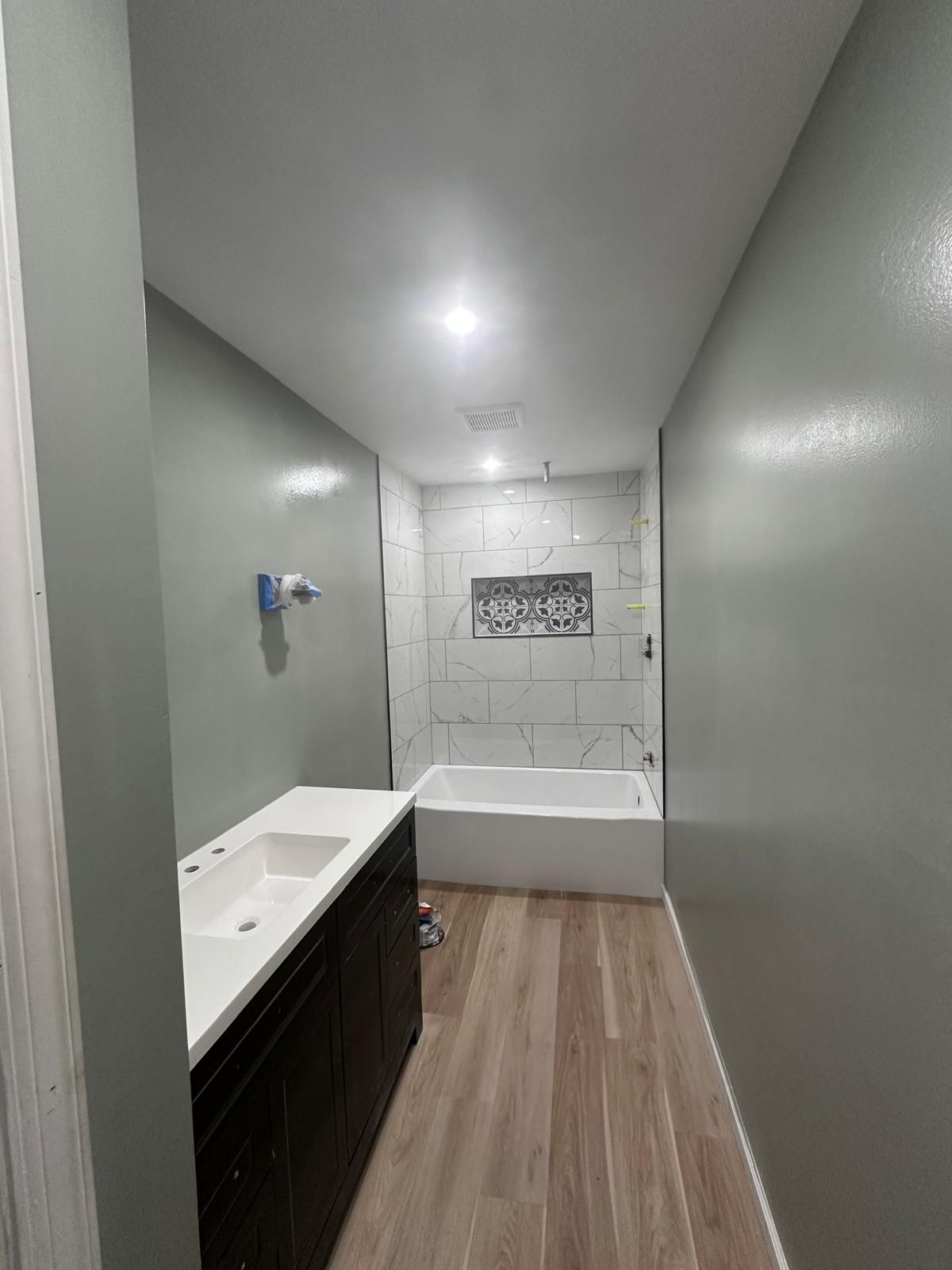 Narrow modern bathroom with vanity on left, bathtub at end, and wood-look flooring.