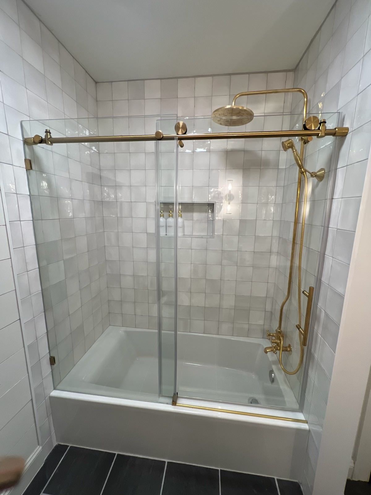 White-tiled bathroom with a glass shower enclosure and gold fixtures over a bathtub.