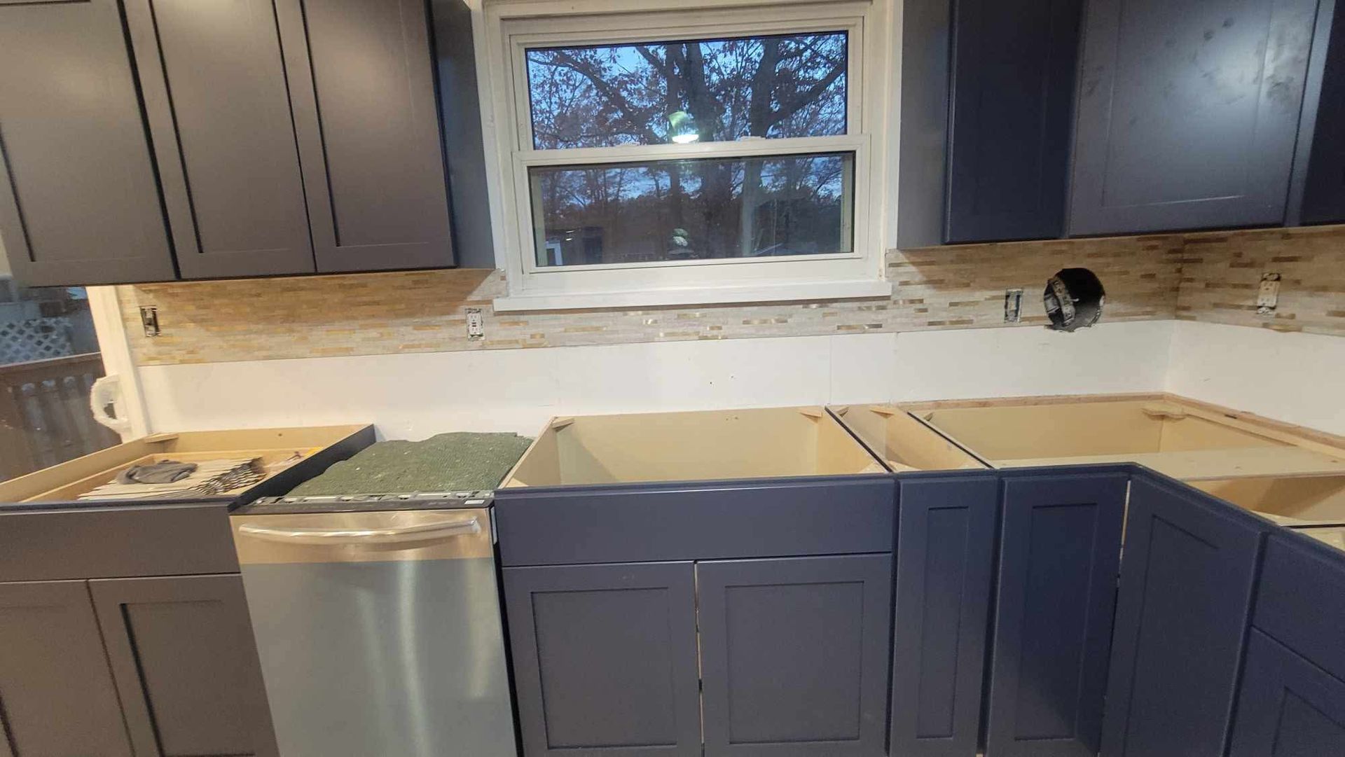 Kitchen under renovation with blue cabinets, uncovered countertops, and a window above the sink.