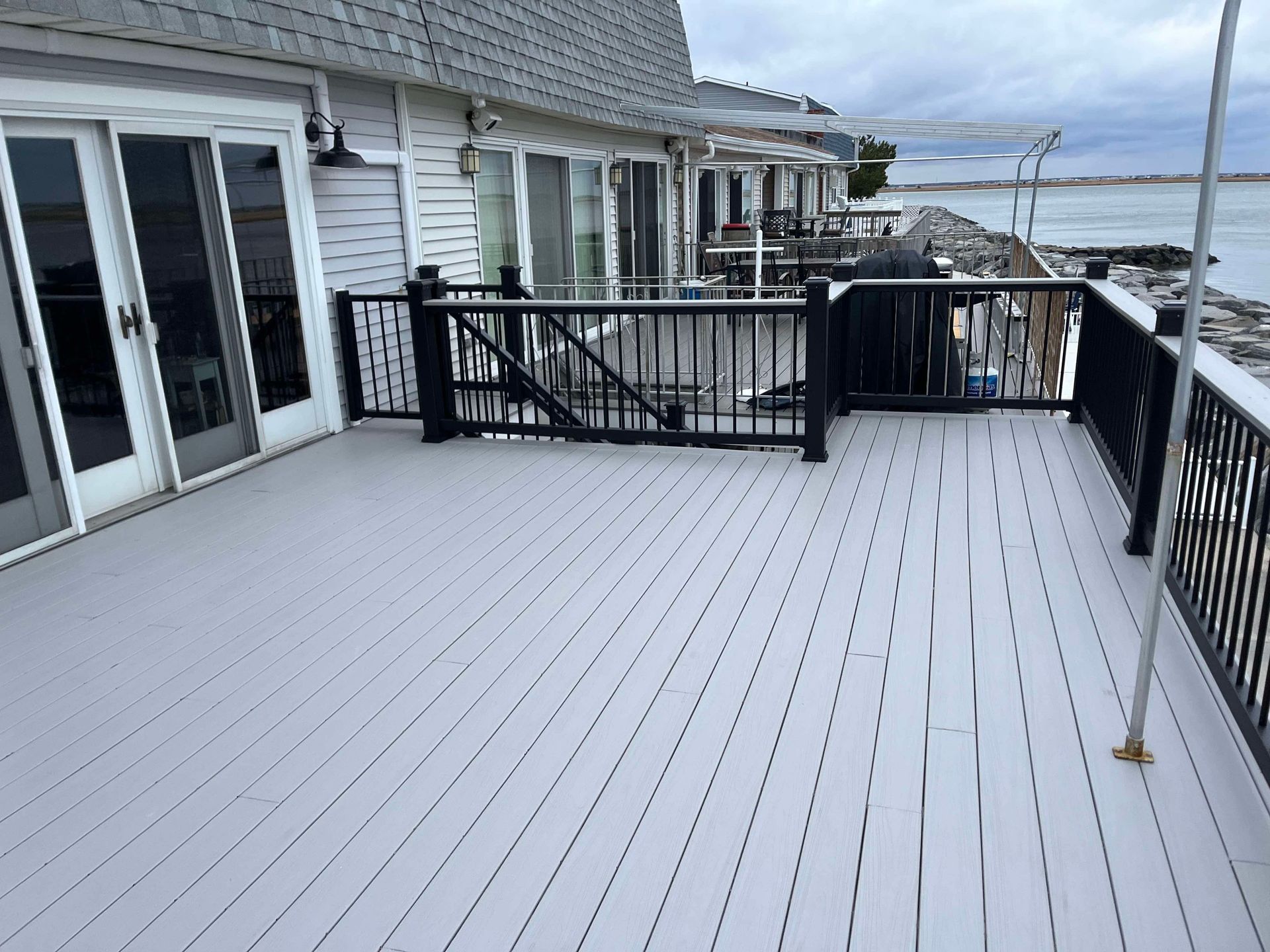 Snow-covered patio deck beside a house, with black railings and water visible beyond.