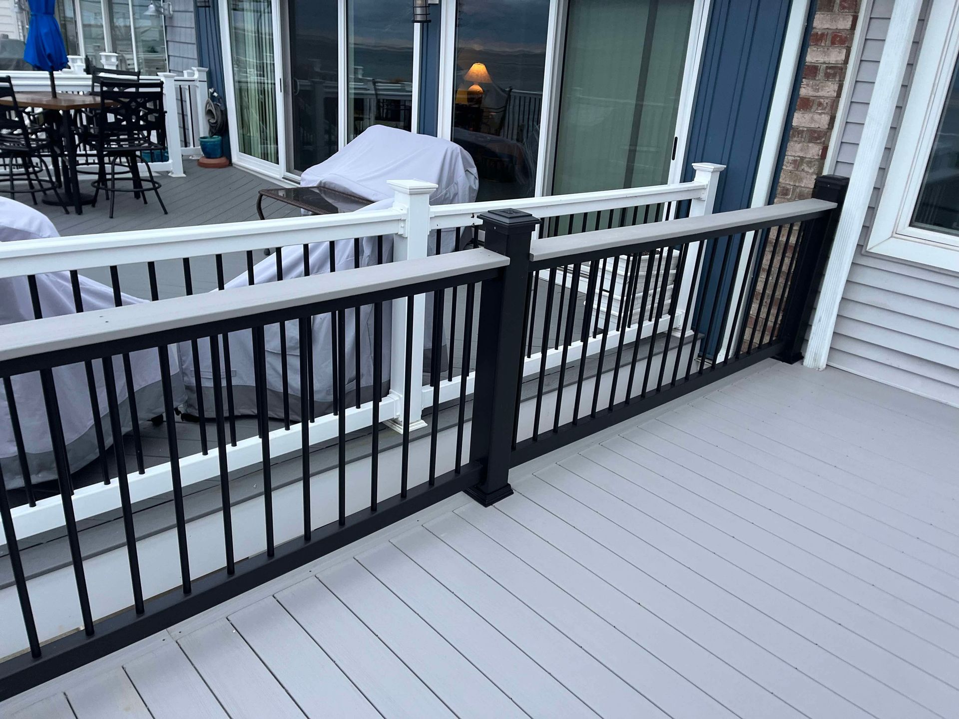 Patio deck with black railing, gray flooring, and a covered grill beside a house window.