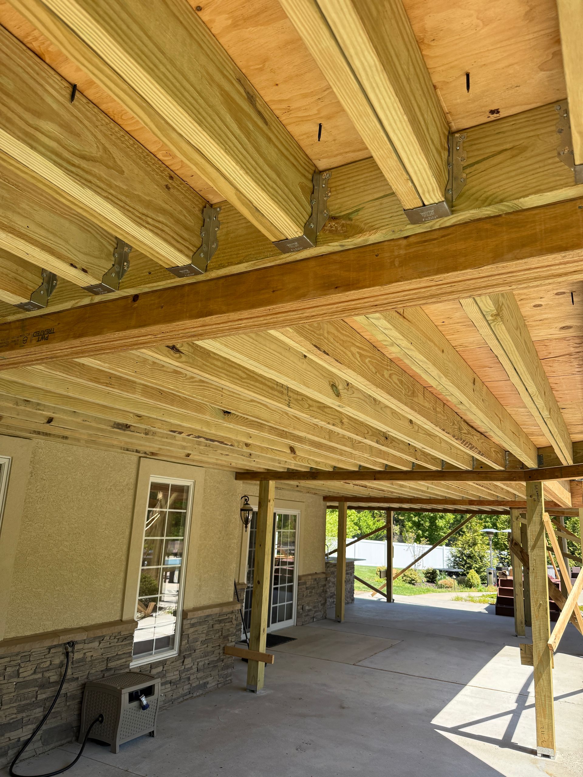 Covered patio under construction with exposed wooden beams and concrete floor, opening to a yard outside