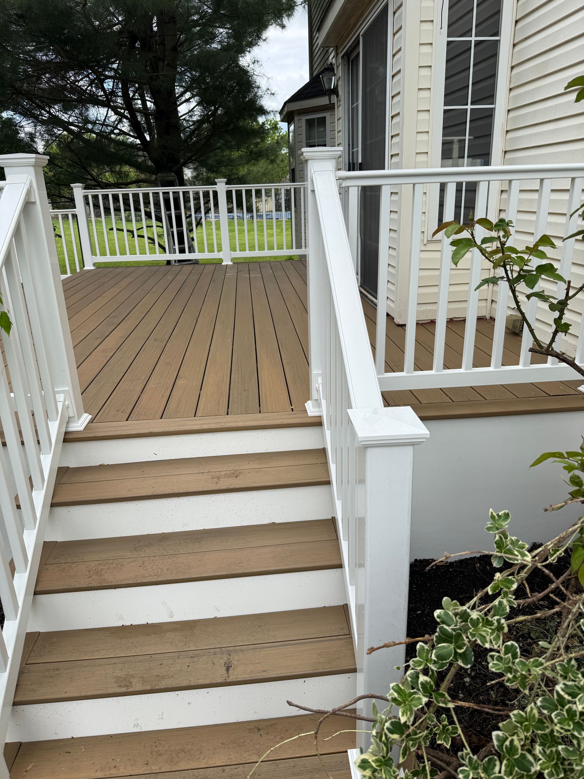White-railed front porch deck with stairs, tan boards, and a house exterior surrounded by trees