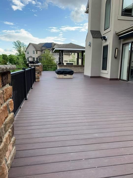 Spacious wooden deck beside a house, with black railing, stone wall, and outdoor seating overlooking neighboring homes.