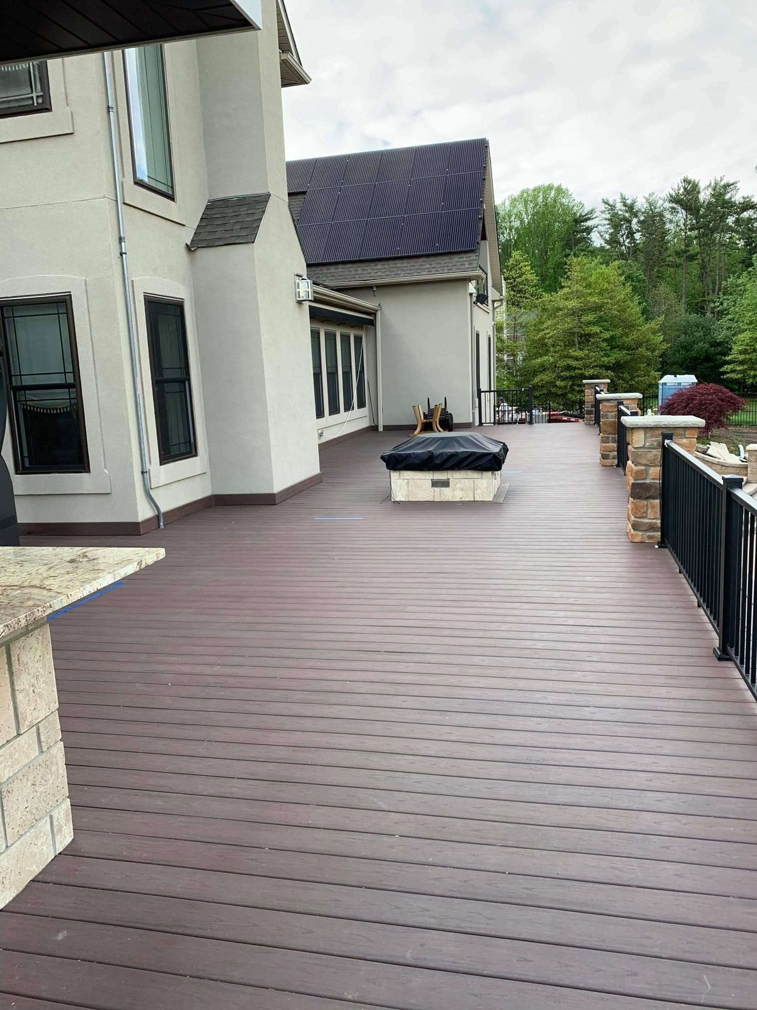 Long wooden deck beside a white house, with potted plants and railing overlooking trees.