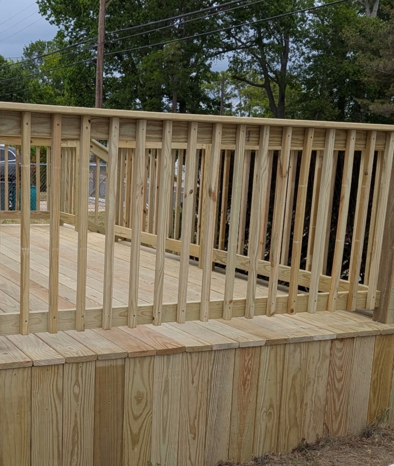 Wooden deck ramp with vertical railings and tan stone base outdoors, with trees in the background