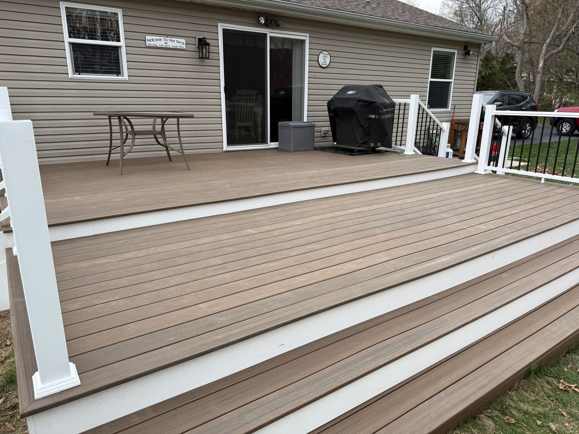 Backyard deck with stairs, sliding door, grill, and patio chairs beside a beige house
