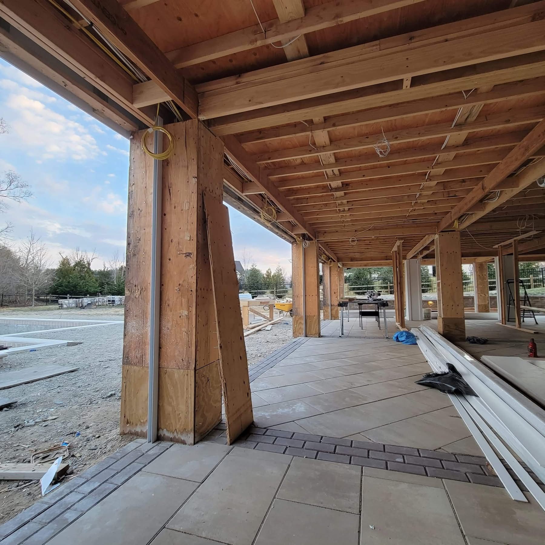 Partially built wooden patio with exposed beams, concrete floor, and stacked lumber at a snowy construction site