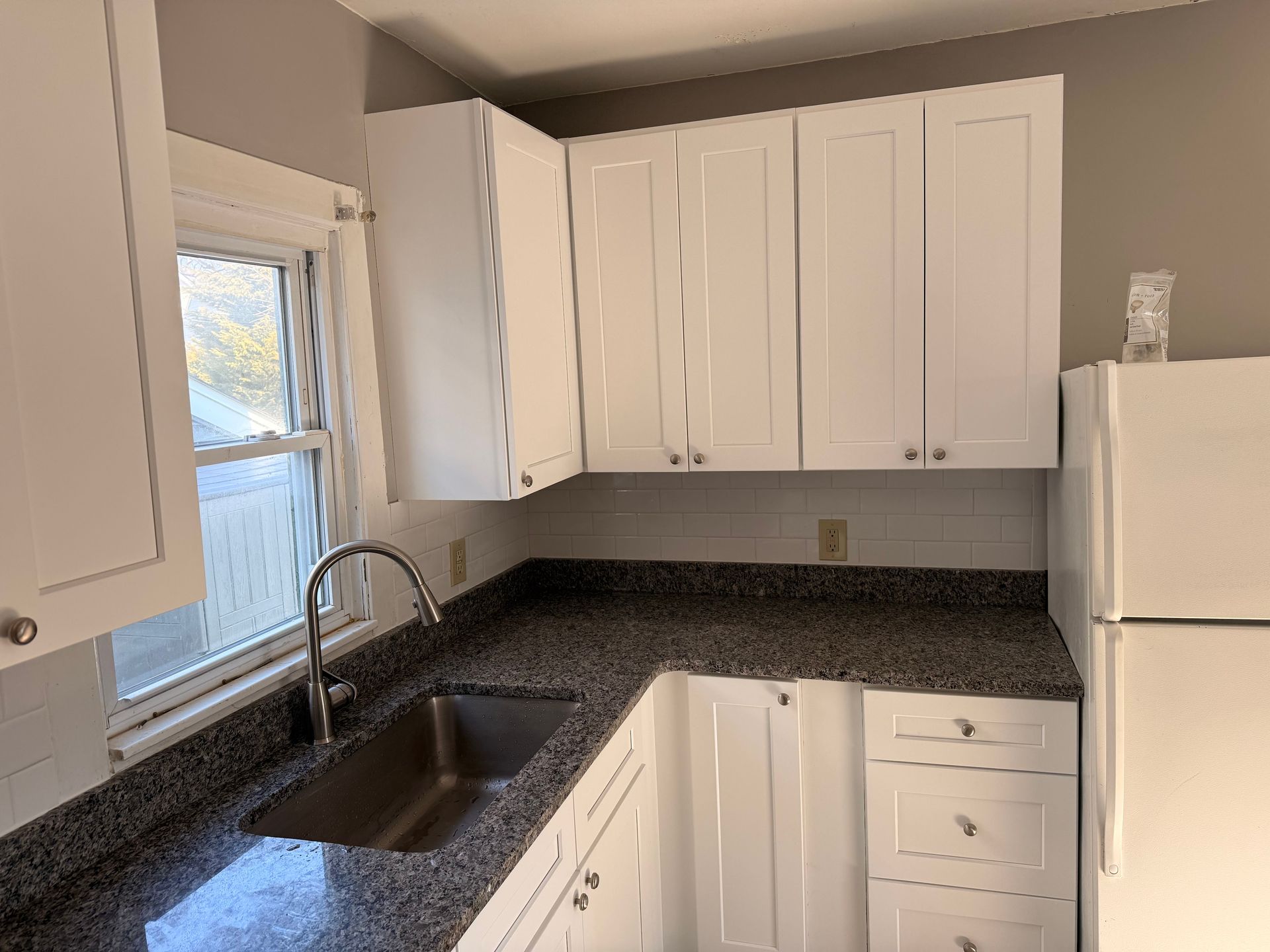 Small white kitchen with granite counters, sink by a window, and a white refrigerator.