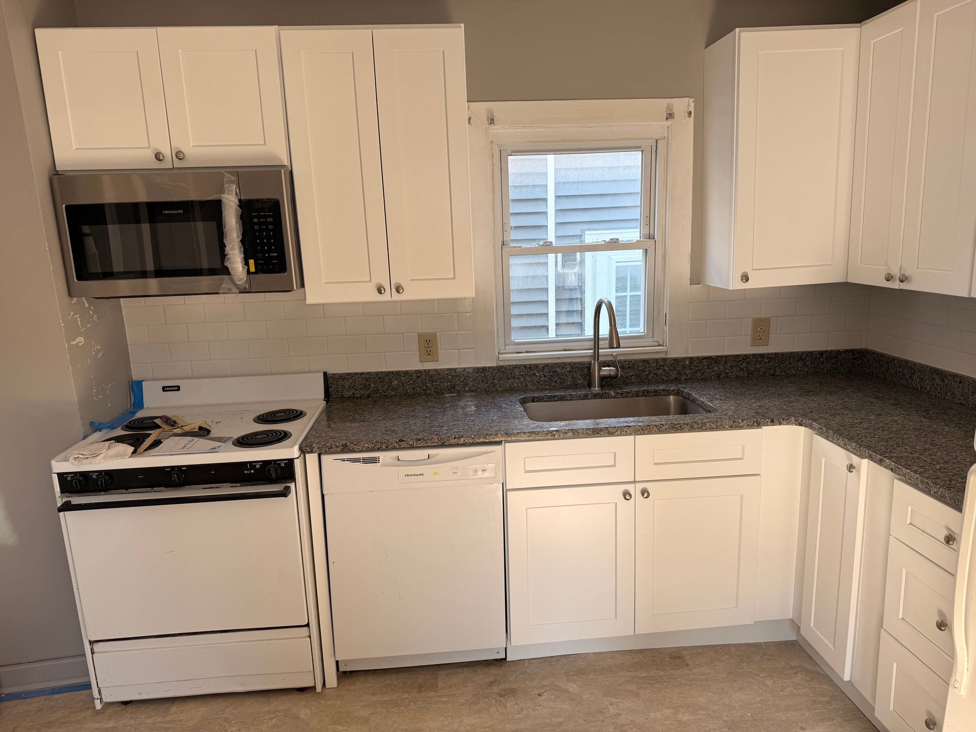 Small white kitchen with stove, microwave, sink under window, and granite countertops