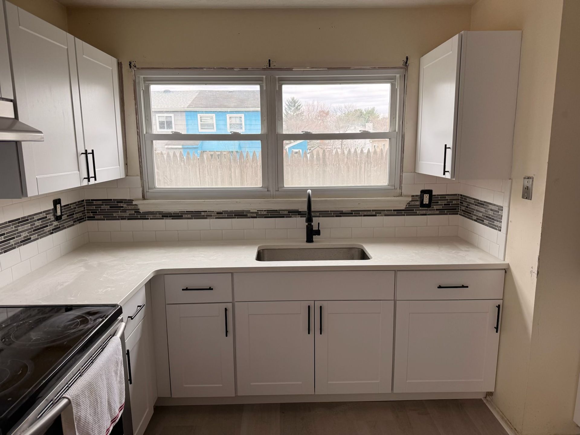 Small kitchen with white cabinets, black countertop, sink under window, and black stove on the left.