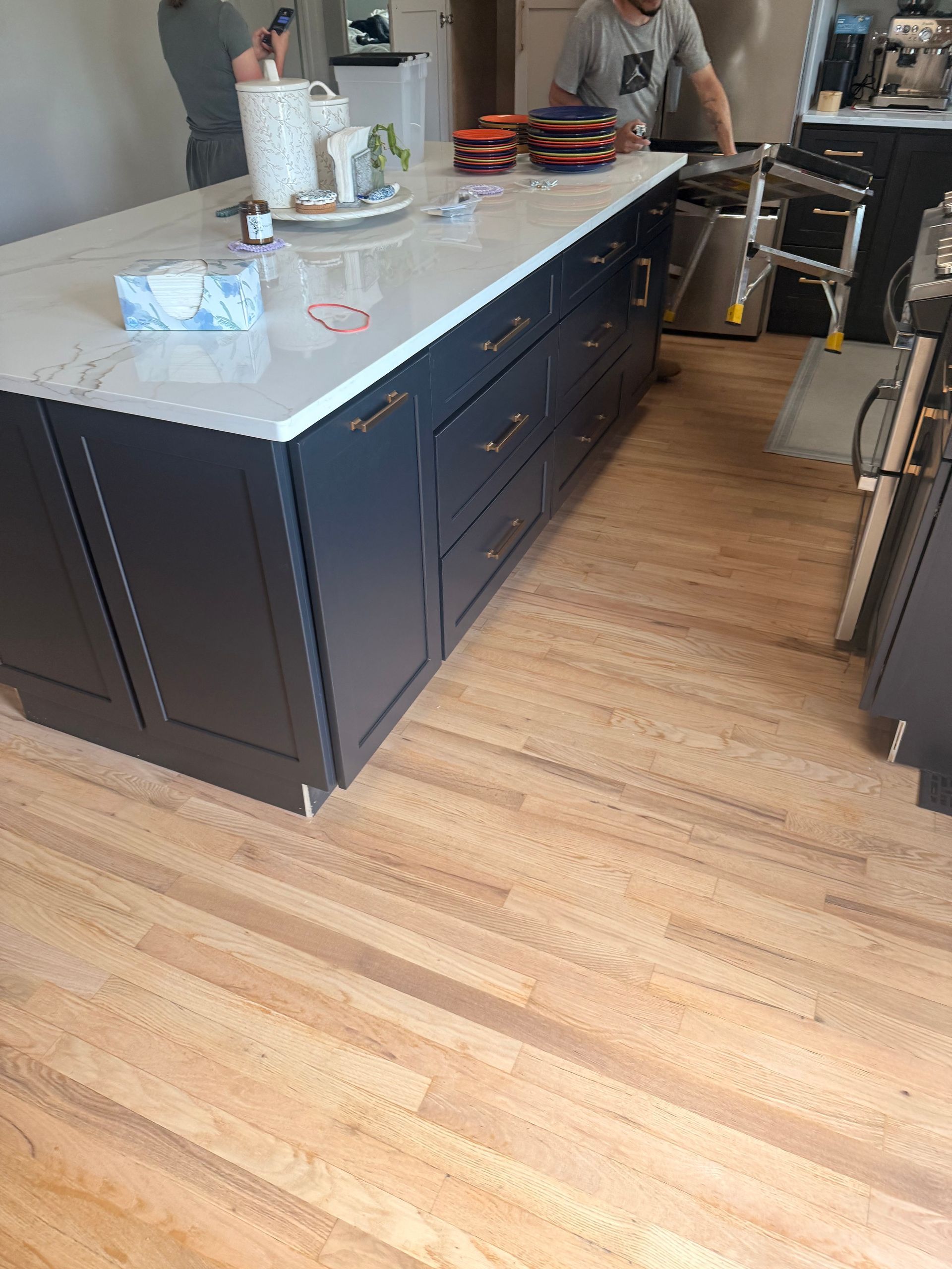 Modern kitchen with dark island cabinets, light wood floor, and two people preparing items on the counter