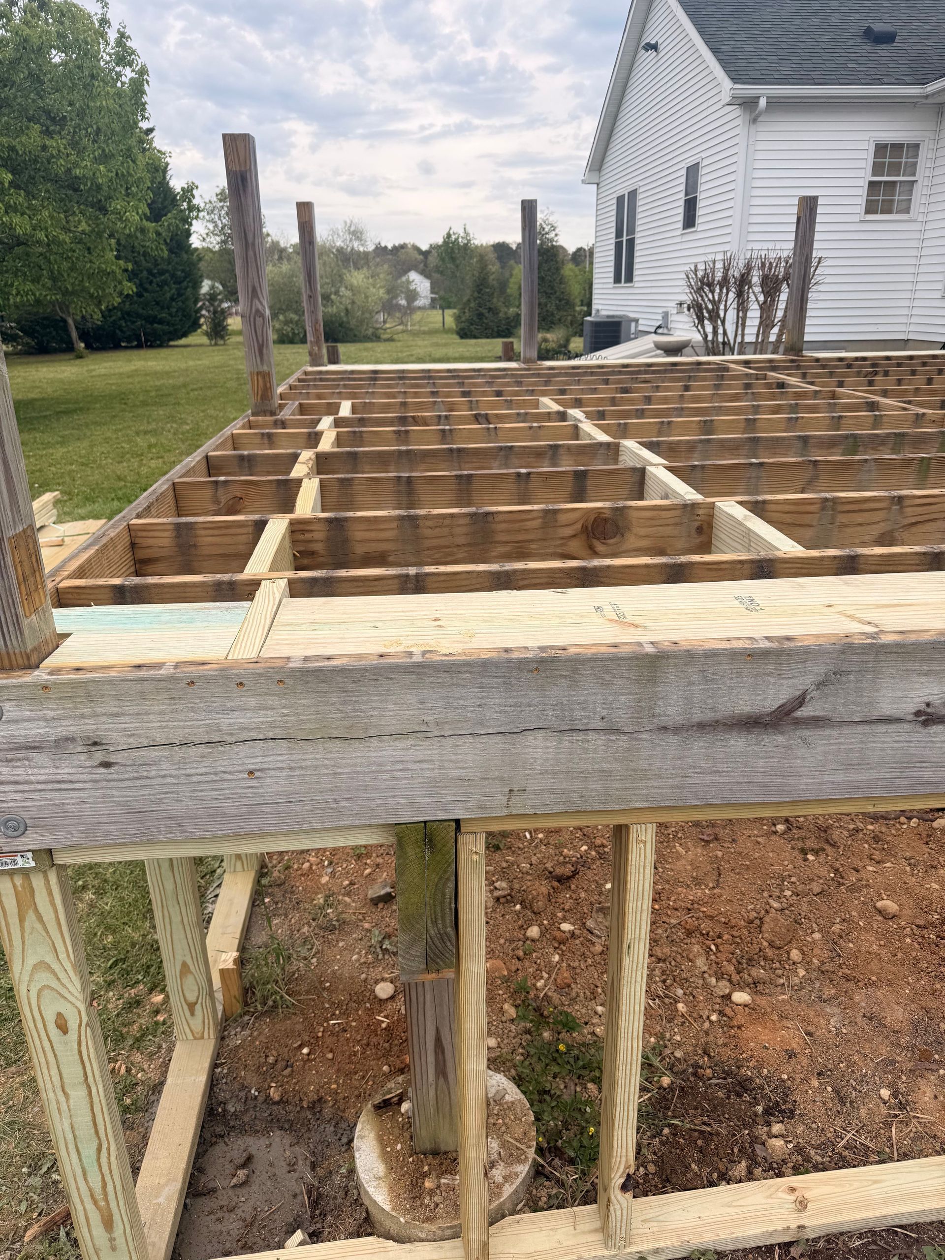 Wooden deck frame under construction beside a white house, with support posts and beams visible.