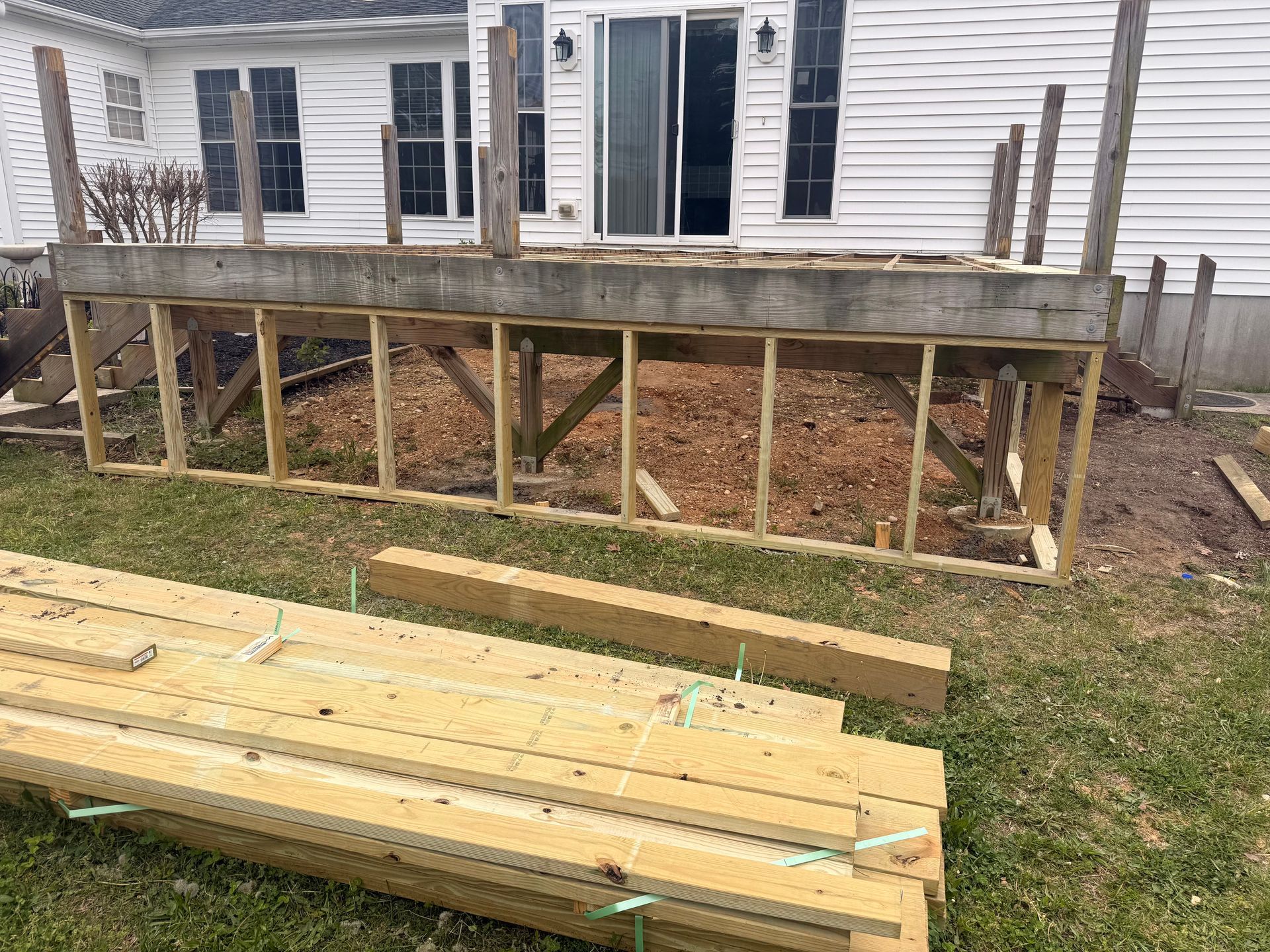 Raised wooden deck under construction beside a white house, with lumber stacked in the yard.