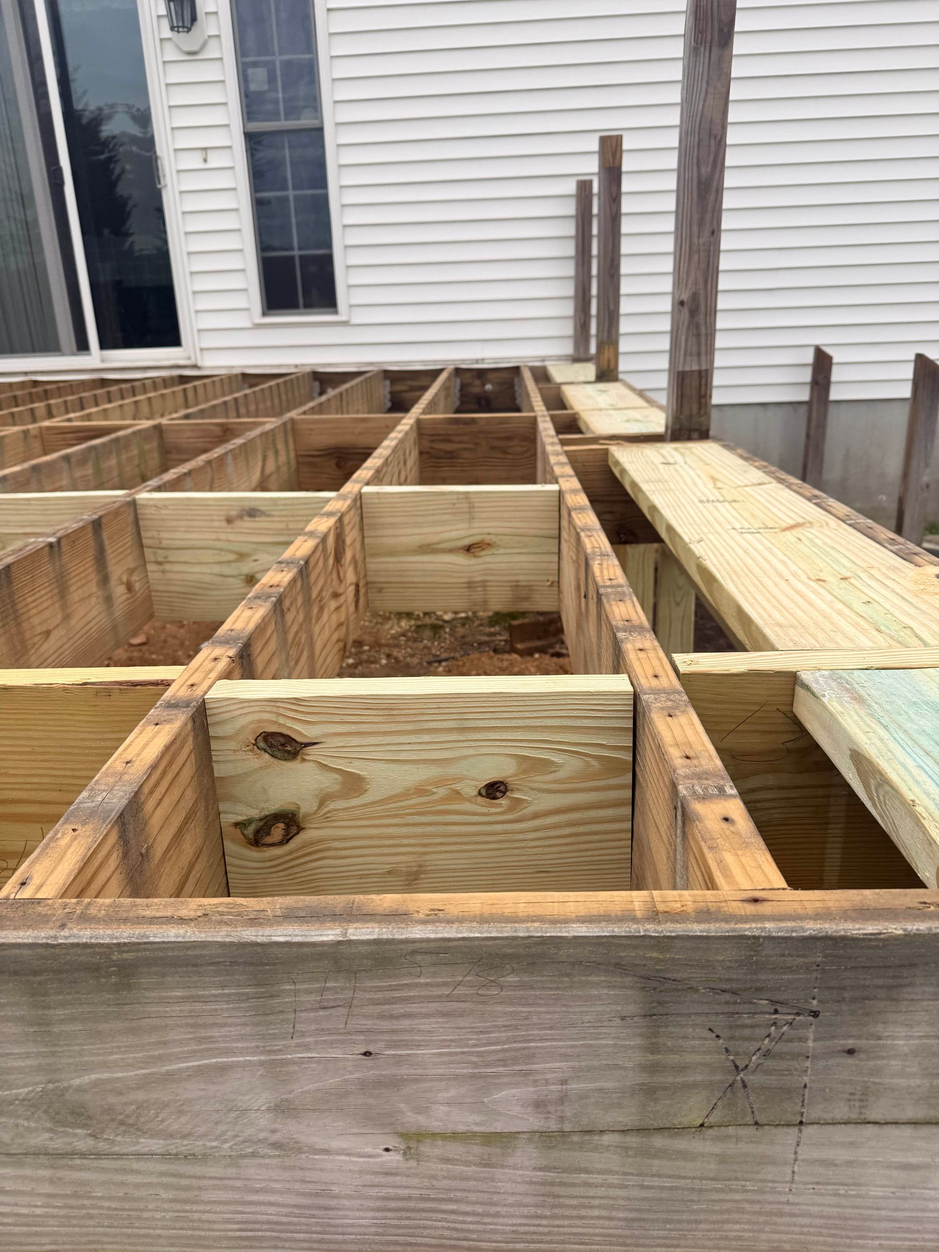 Partially built wooden deck framing beside a white house with windows and posts