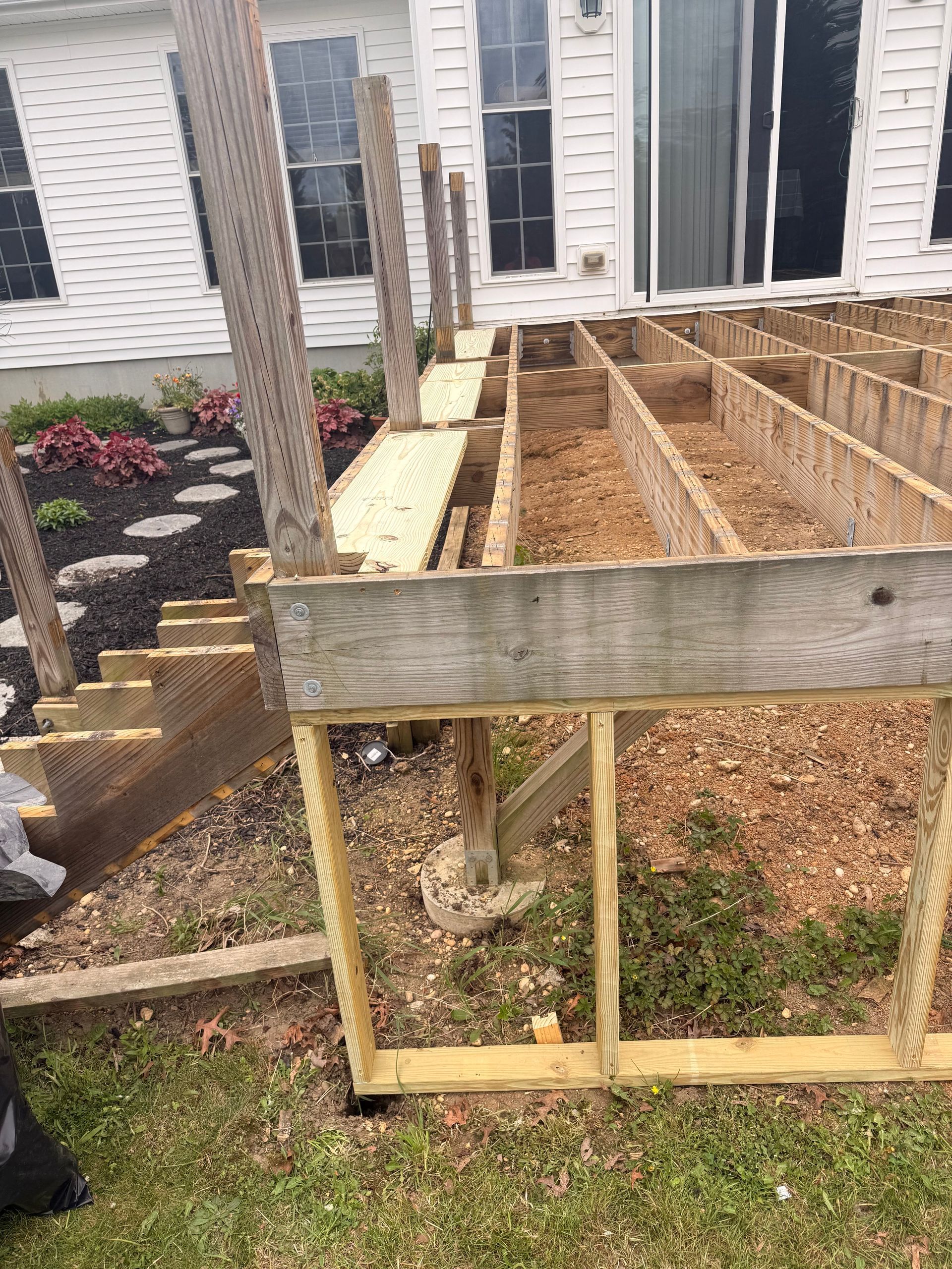 Raised garden bed frame with wooden boards and support posts beside a house and walkway