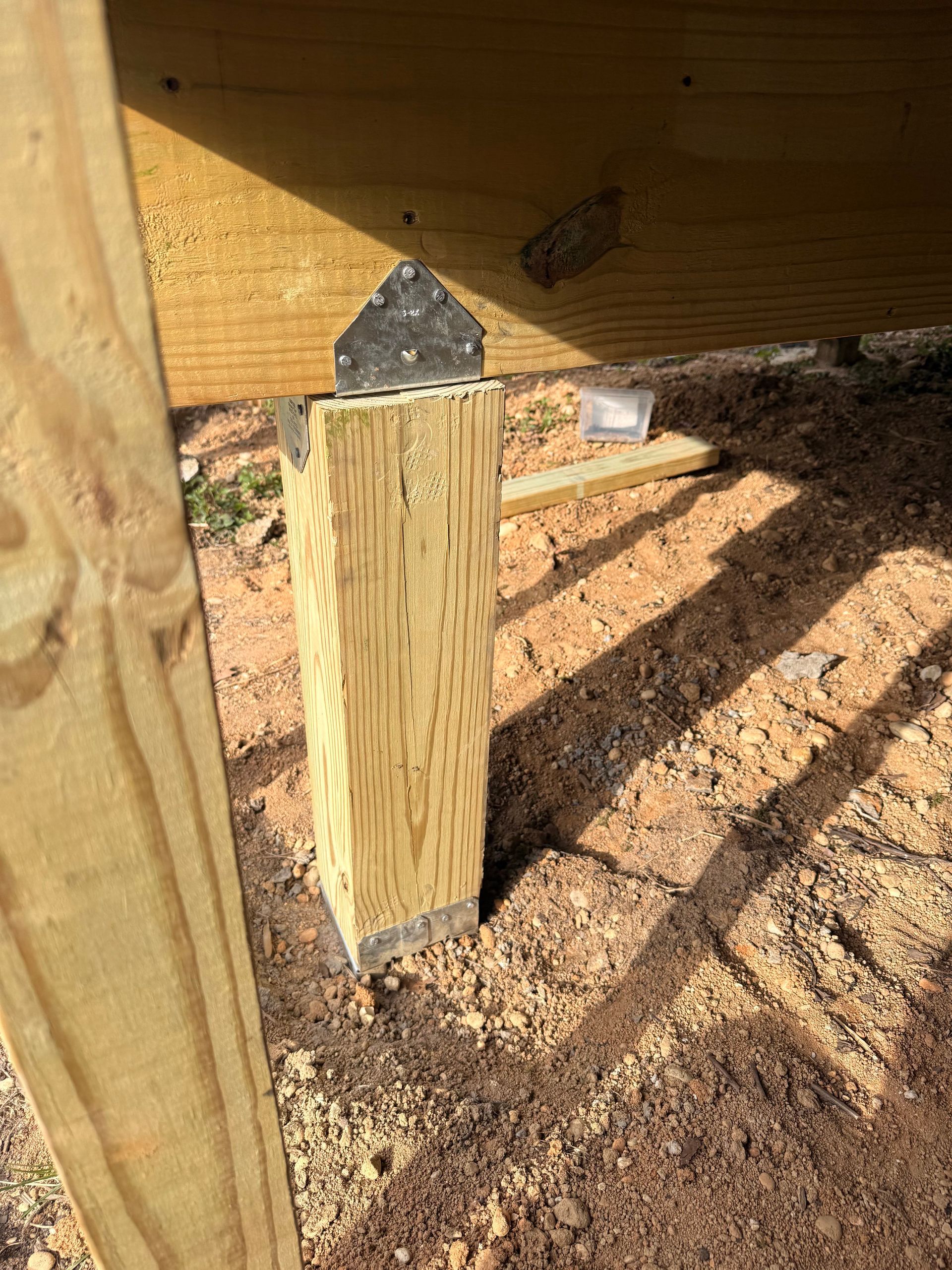 Wooden support post under a deck, with a metal bracket at the top on a dirt foundation.