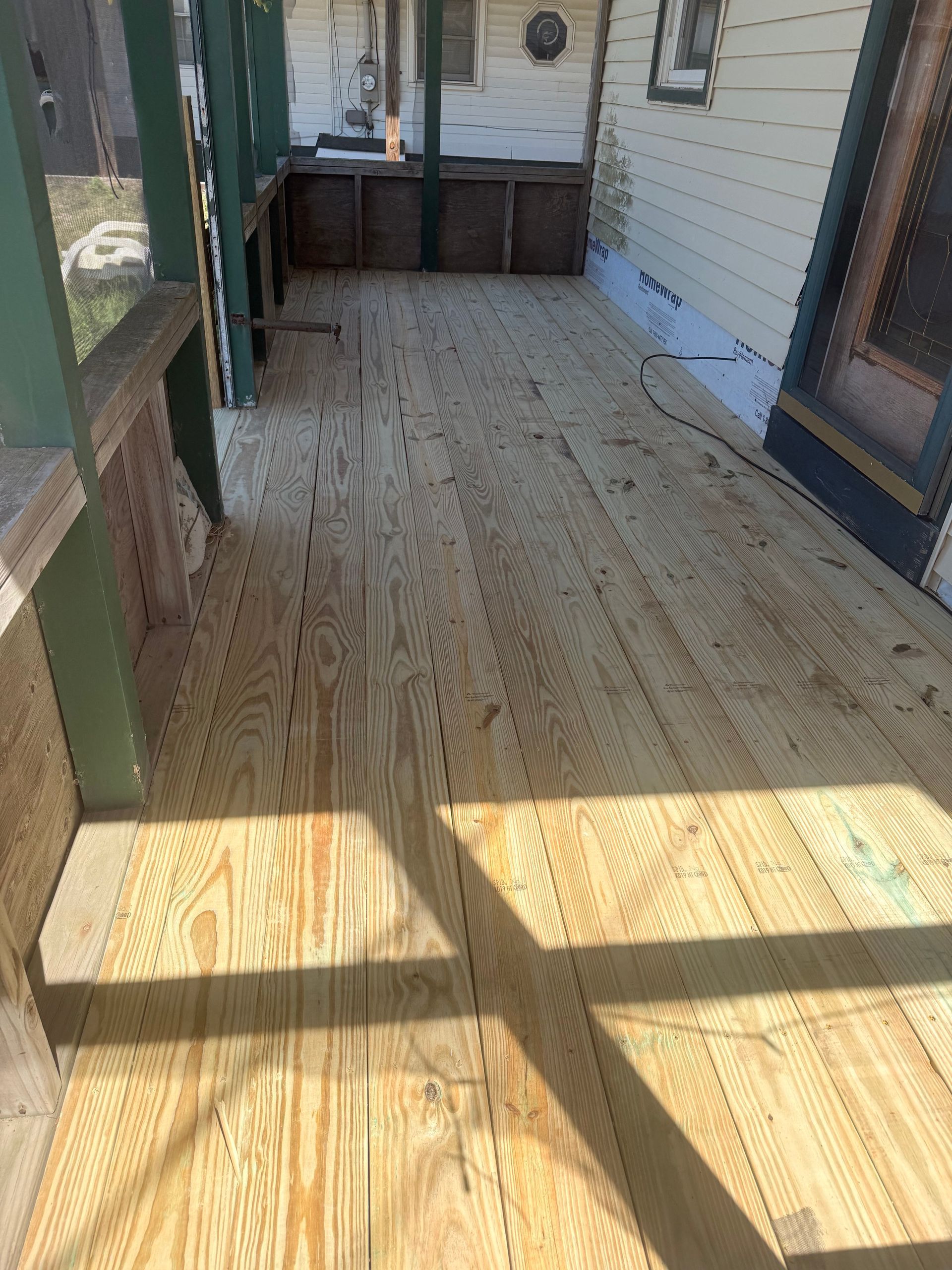 Empty wooden porch/deck with railings and sunlight casting shadows, beside a house.